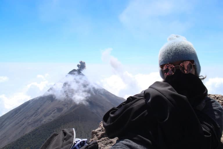 A woman wearing a jacket, sunglasses, and a knit hat faces the camera at the summit of Acatenango while a volcano rises in the background with a small explosive puff of ash emerging from the top.