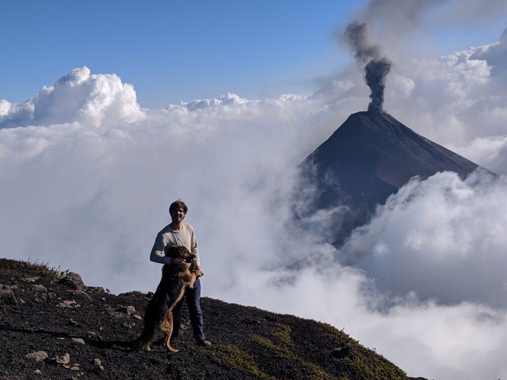 A young man and a German shepherd stand together on a dark ridge with a volcano erupting clouds of grey ash in the background.