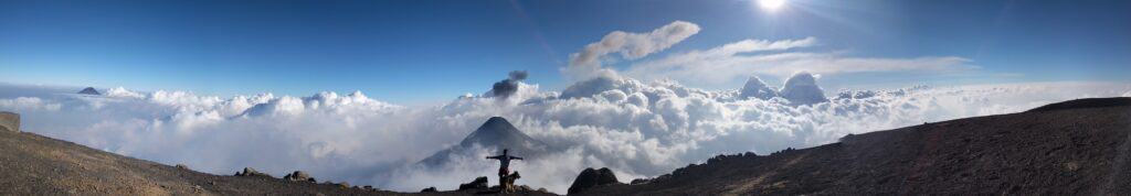 A man raises his arms triumphantly while his German Shepherd walks beside him. In the background is a sea of fluffy clouds, lit by  a white sun, which a volcano rising from their midst.