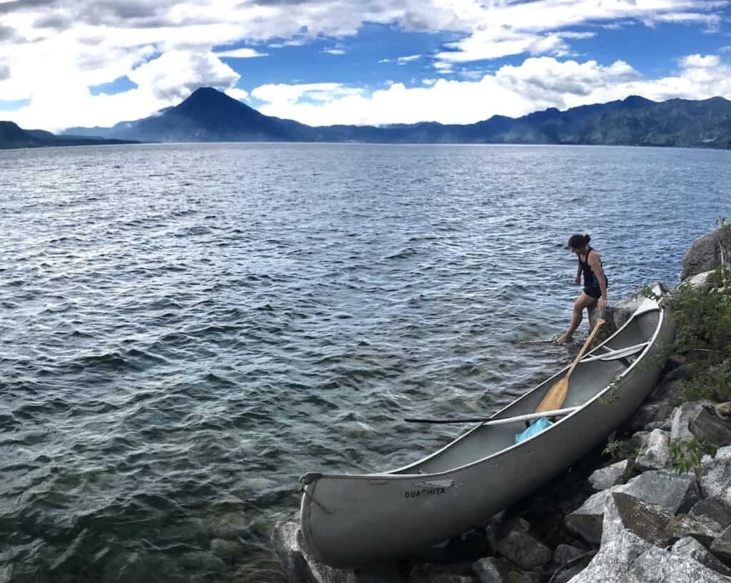 La imagen muestra el lago de Atitlán, con aguas oscuras y onduladas y un volcán que se eleva azul y oscuro sobre el agua. En primer plano hay una canoa tirada sobre las rocas y una mujer vadeando el lago.