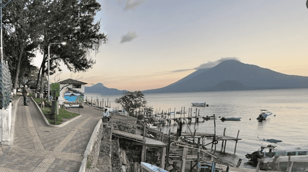 La imagen muestra la orilla lejana del lago de Atitlán, salpicada de volcanes, con un camino en primer plano que se bifurca a izquierda y derecha. En el centro hay un edificio redondo pintado con un mural del lago entre los brazos de una mujer.