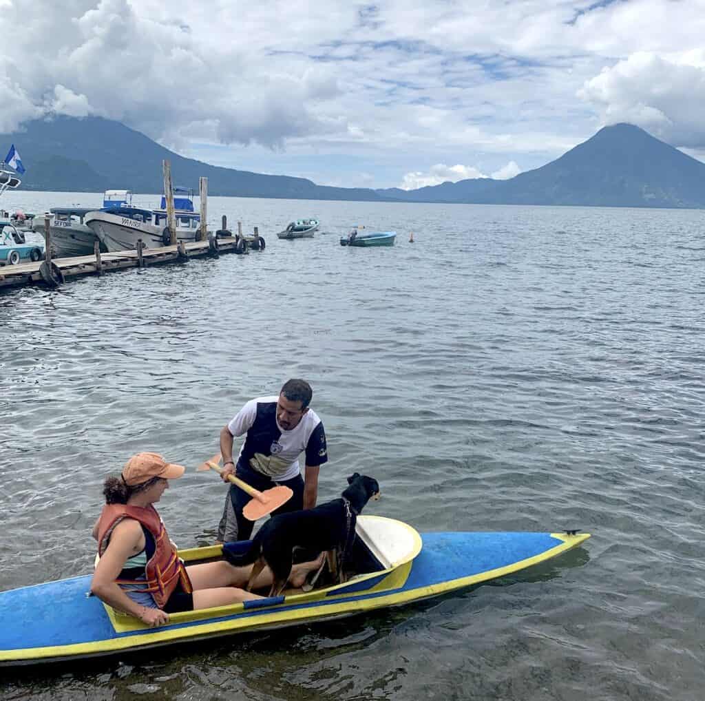 Una mujer y su perro se colocan dentro de un kayak en el lago de Atitlán, mientras un hombre lo mantiene firme y le entrega el remo. Hay espacio adicional para el perro delante del palista. Los volcanes son visibles al otro lado del lago.