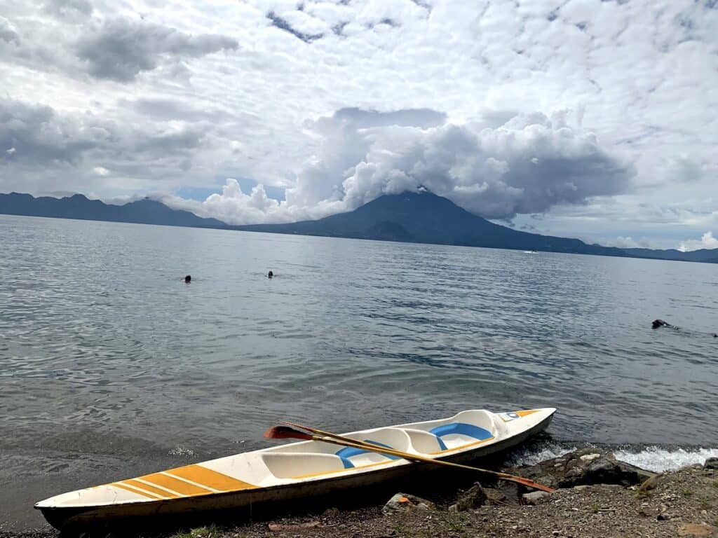 Un kayak de rayas amarillas yace cruzado a la orilla de un lago. Al otro lado del agua, dos volcanes se alzan picudos y azules contra la orilla, mientras el cielo se llena de nubes esponjosas y escamosas.