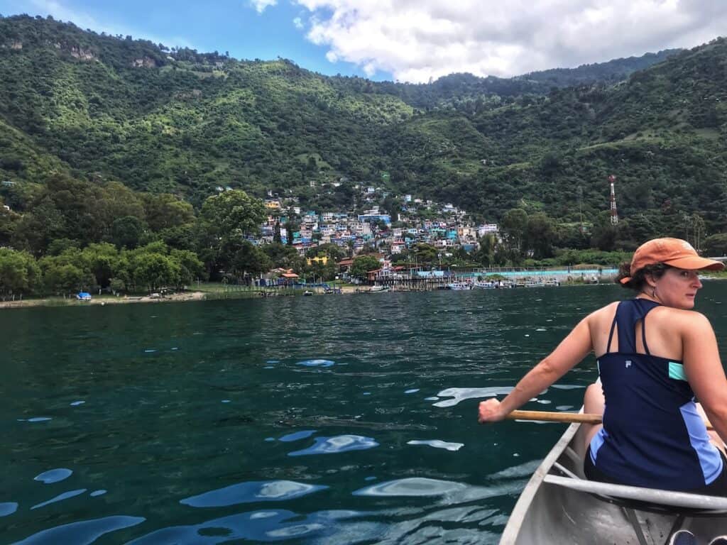 A woman in a canoe looks to the side in the foreground while the background shows a green tree-clad ridge, nestling a colorful town that rises from a reedy shore.