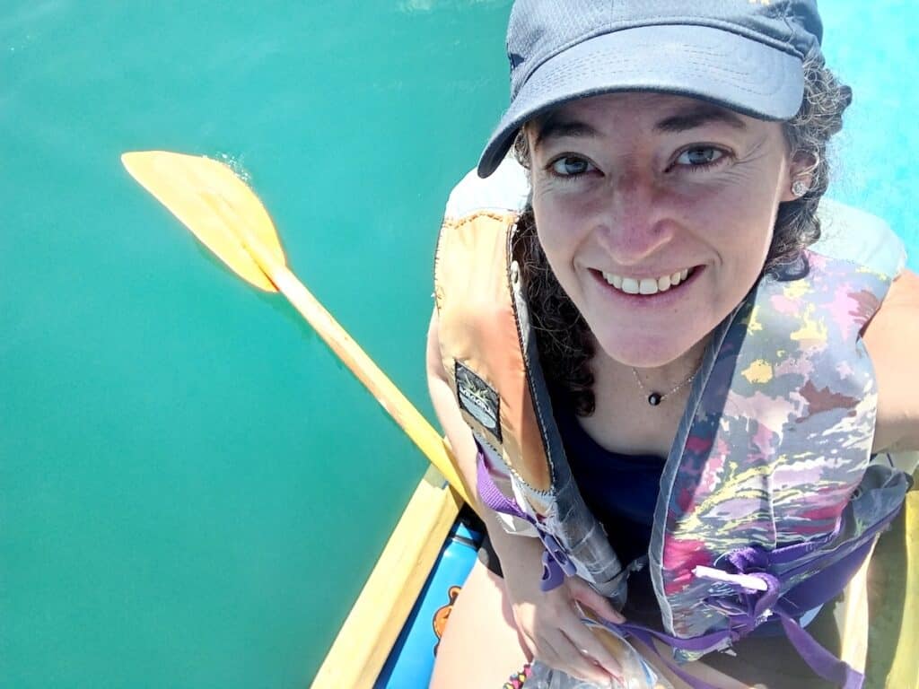 A woman wearing a hat and lifejacket sits in a kayak overtop tranquil turquoise water. Besides the kayak, you can just see the edge of her water bottle and a plastic bag with her phone.