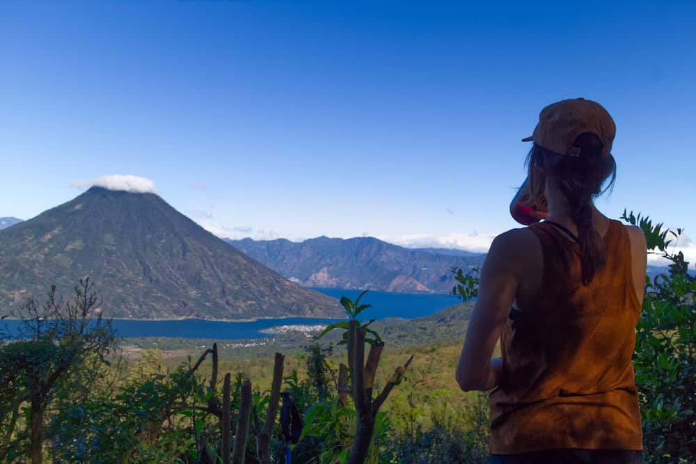 A woman in a baseball cap drinks water, enjoying views of a volcano rising from a lake.