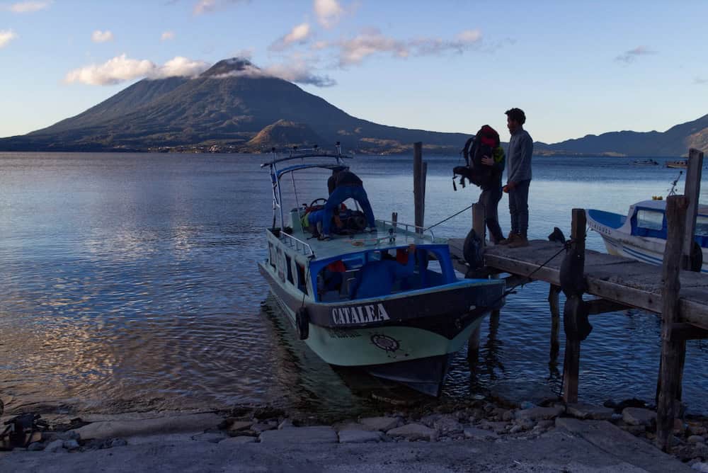 Tolimán and Atitlán volcanoes rise above Lake Atitlán, painted golden in the dawn light. Small puffy clouds trail across the sky. In the foreground, men load a hiking backpack onto a small speedboat.