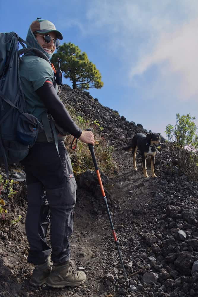 A man wearing a hiking backpack, hat, and sunglasses grins at the camera. The trail leads past him, across a 45 degree slope. The path is black and sandy, and surrounded by volcanic rocks. 