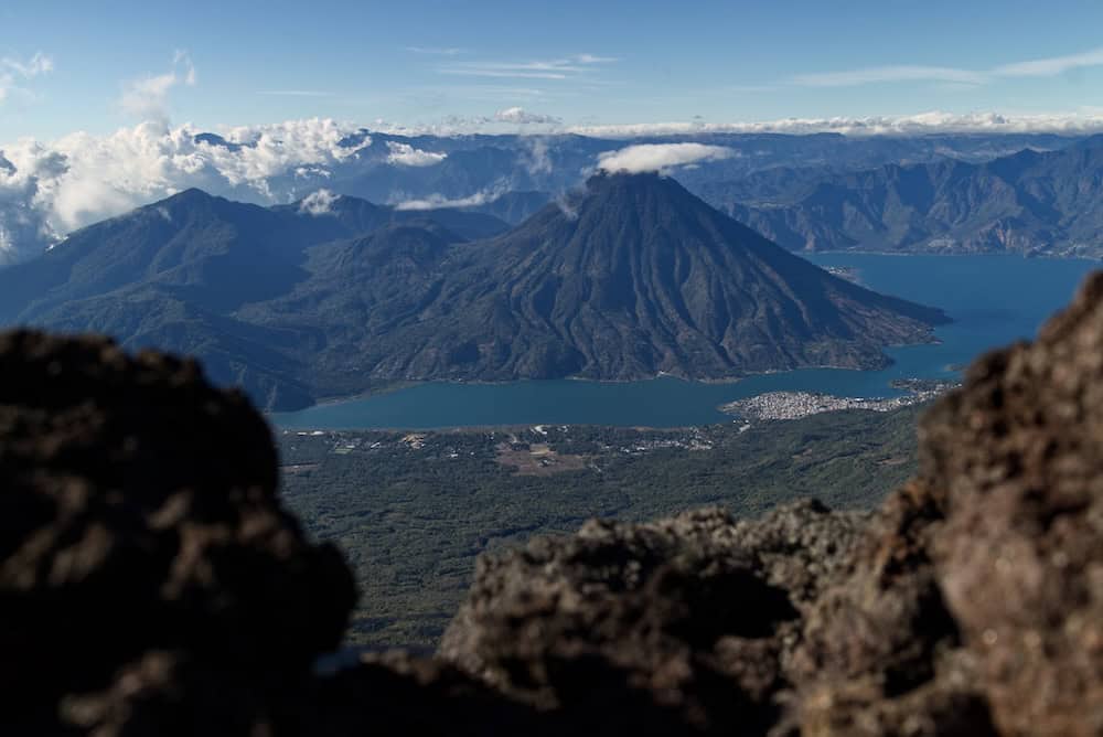 A volcano rises above a lake far below, framed between rugged volcanic rocks.