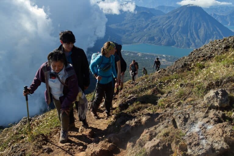 Unos excursionistas suben por una vereda de tierra hacia la cima del volcán Atitlán. Más abajo, se ve un lago azul y las espectaculares crestas de otro volcán.