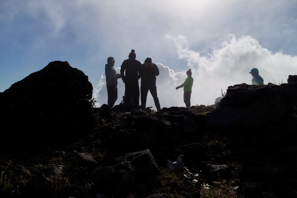 A group of hikers are silhouetted between a large boulder and a camping windbreak made of rocks.