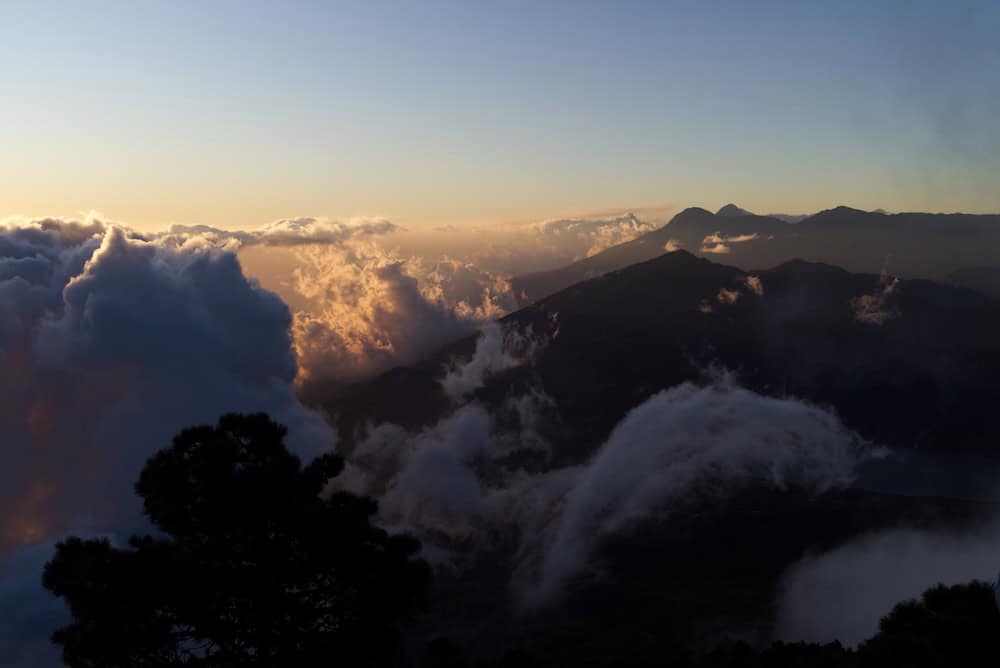 Clouds gleam gold where the setting sun catches them. Volcanoes parade into the distance below, their peaks a shadowed blue against the sky.