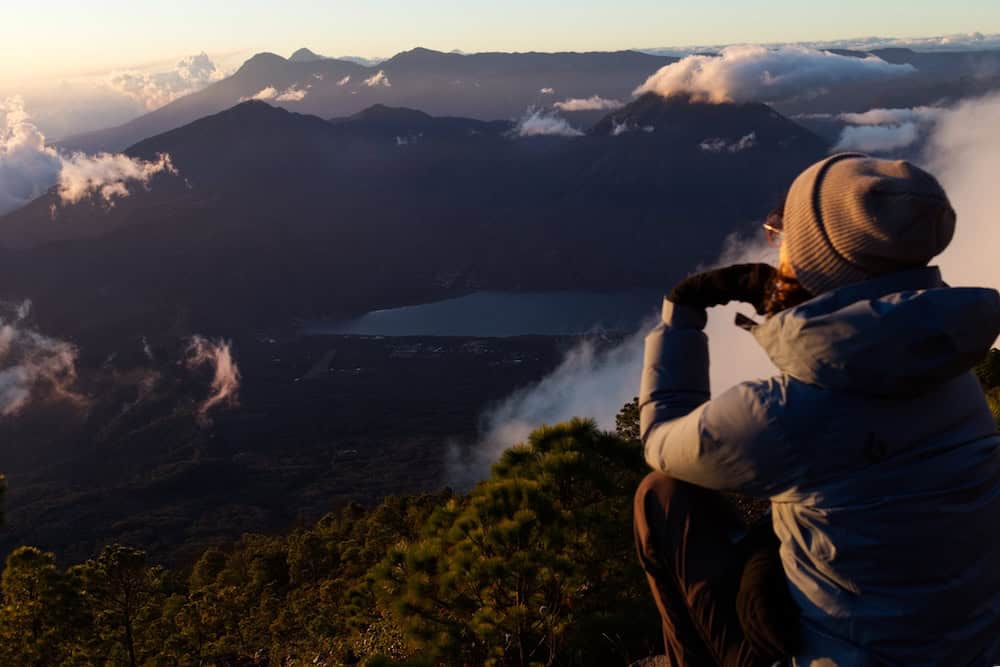 A woman hiker sits on the side of a trail while the sun sets. The clouds are white with glints of gold, and the landscape is deep blue. Six volcanoes parade into the distance.