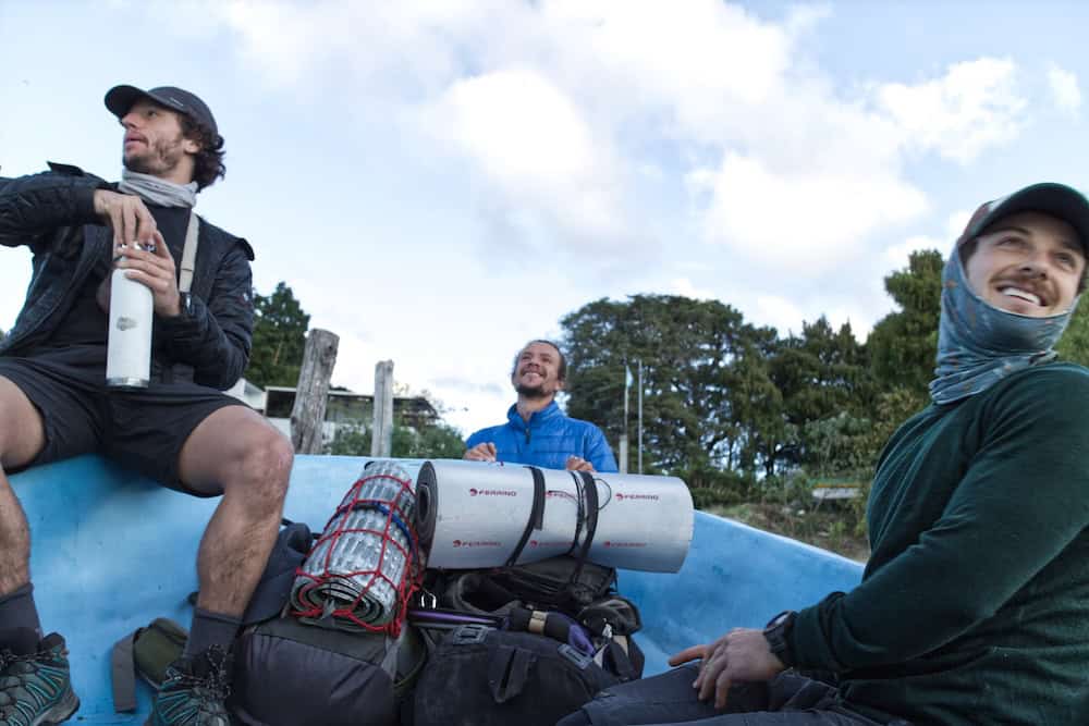 Two young men sit on either side of a speedboat prow, with hiking backpacks between them. A third man stands outside the boat. They are all smiling and looking in different directions.