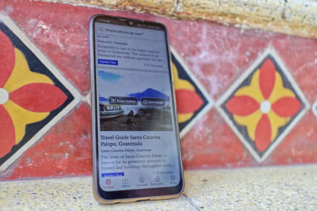 The image shows a phone against a background of red-patterned tiles in a Spanish colonial style. On the phone are travel articles available for use offline.