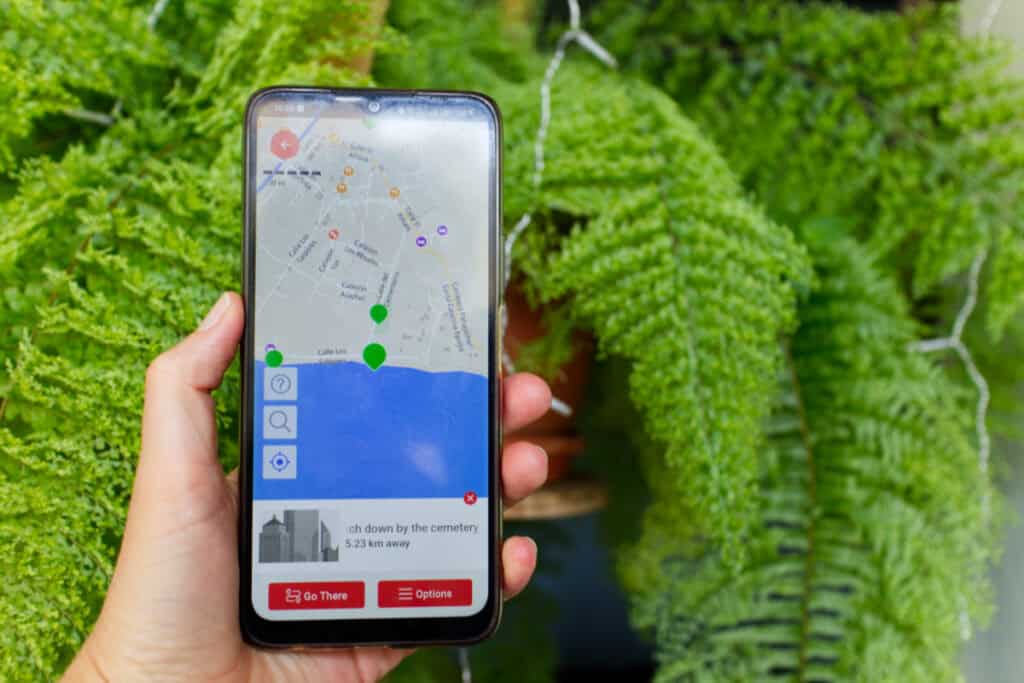 A person holds a phone in front of a bright green fern. On the screen is a map with green location icons.